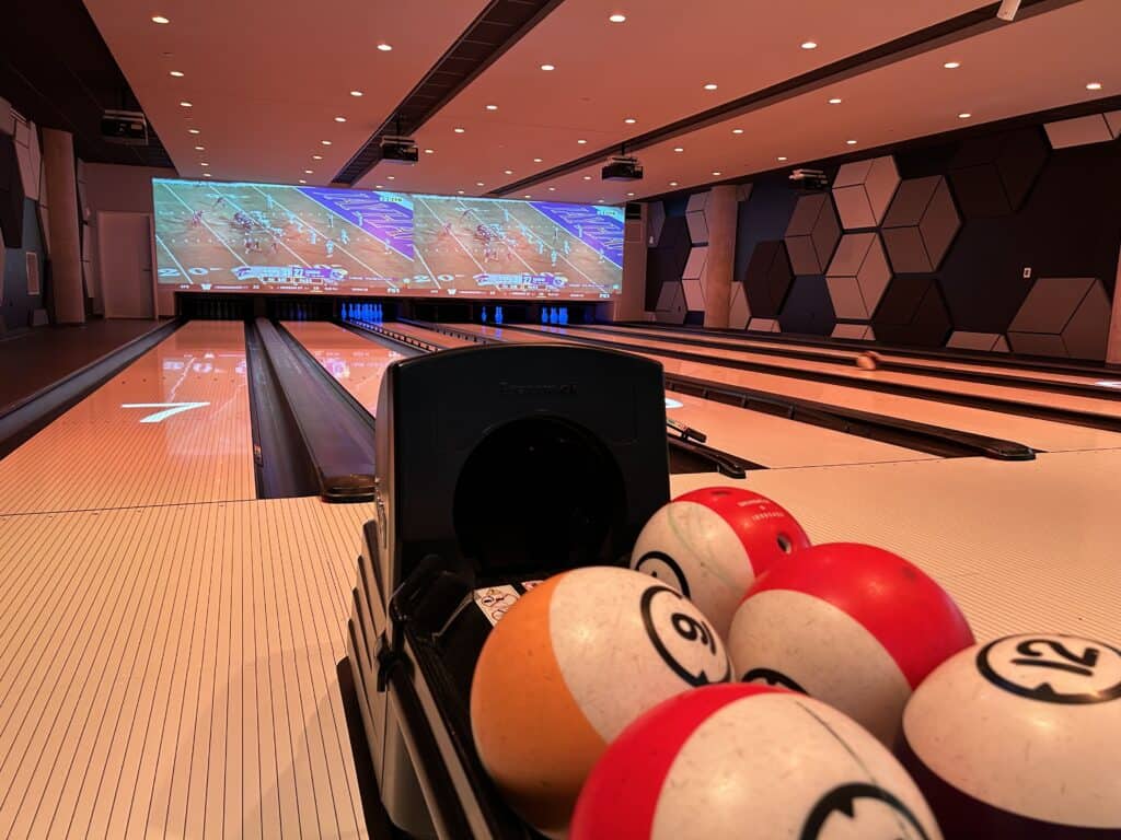 Empty bowling alley showing five lanes, with colorful bowling balls in forefront and large screens showing a football game in distance above bowling pins.